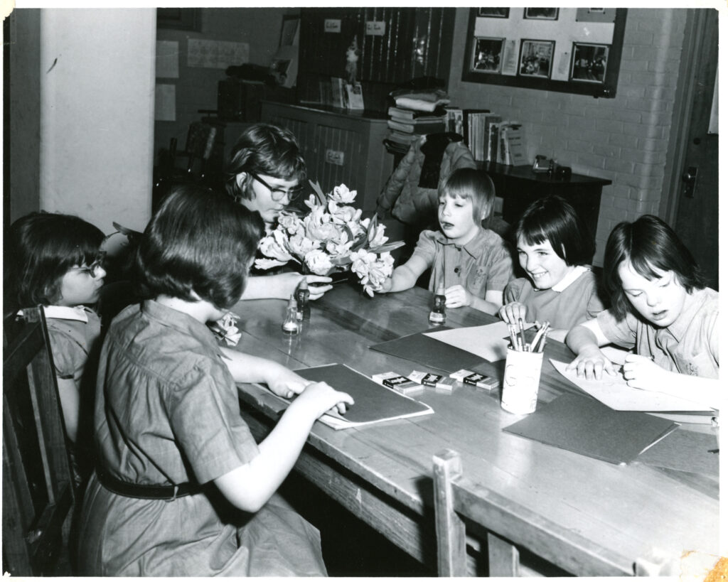Black and white photo of six girls sitting around a table, all wearing Camp Fire Girls uniforms, with papers and pencils. One is holding a bouquet of flowers.