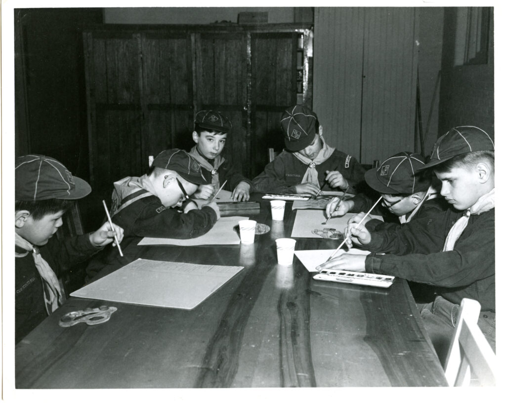 Black and white picture of six boys sitting around a table dressed in boy scouts uniforms, using watercolor paints on pieces of paper.