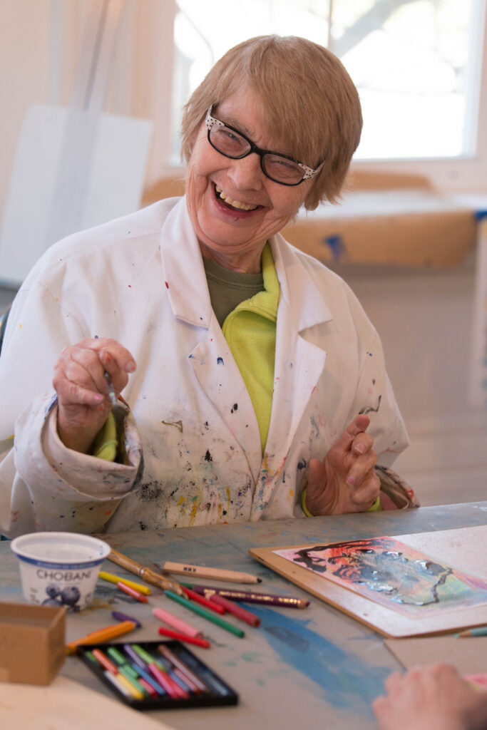 Grace McKenna sits at a table wearing a white smock, painting, and smiling at the camera