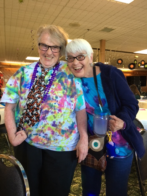 Two older ladies wearing bright colors and grinning at the camera, one with her arm around the other