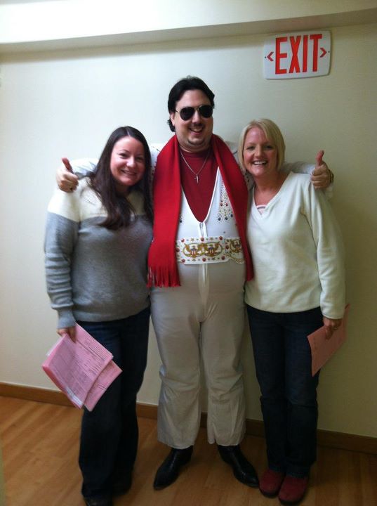 Marco, dressed as Elvis Presley in a white jumpsuit, red shirt and scarf and sunglasses, stands with his arms around two women, giving a double thumbs up