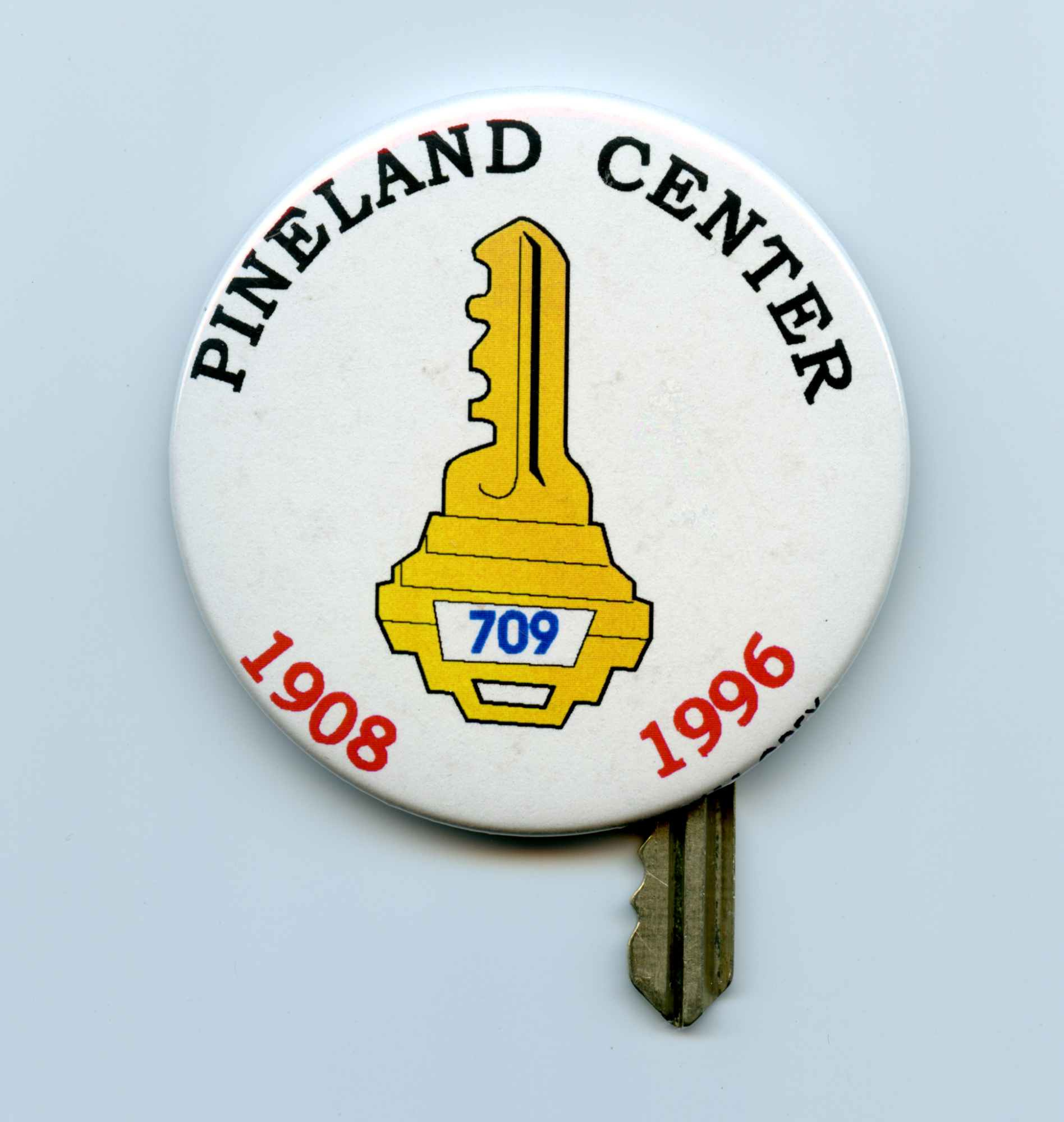 A photo of a round pinback slogan button, with the words “PINELAND CENTER” around the top edge in black, the dates “1908” and “1996” around the bottom edge in red, and a drawing of a yellow key with the number “709” on it. Peeking out from under the button is the bottom of a house key.