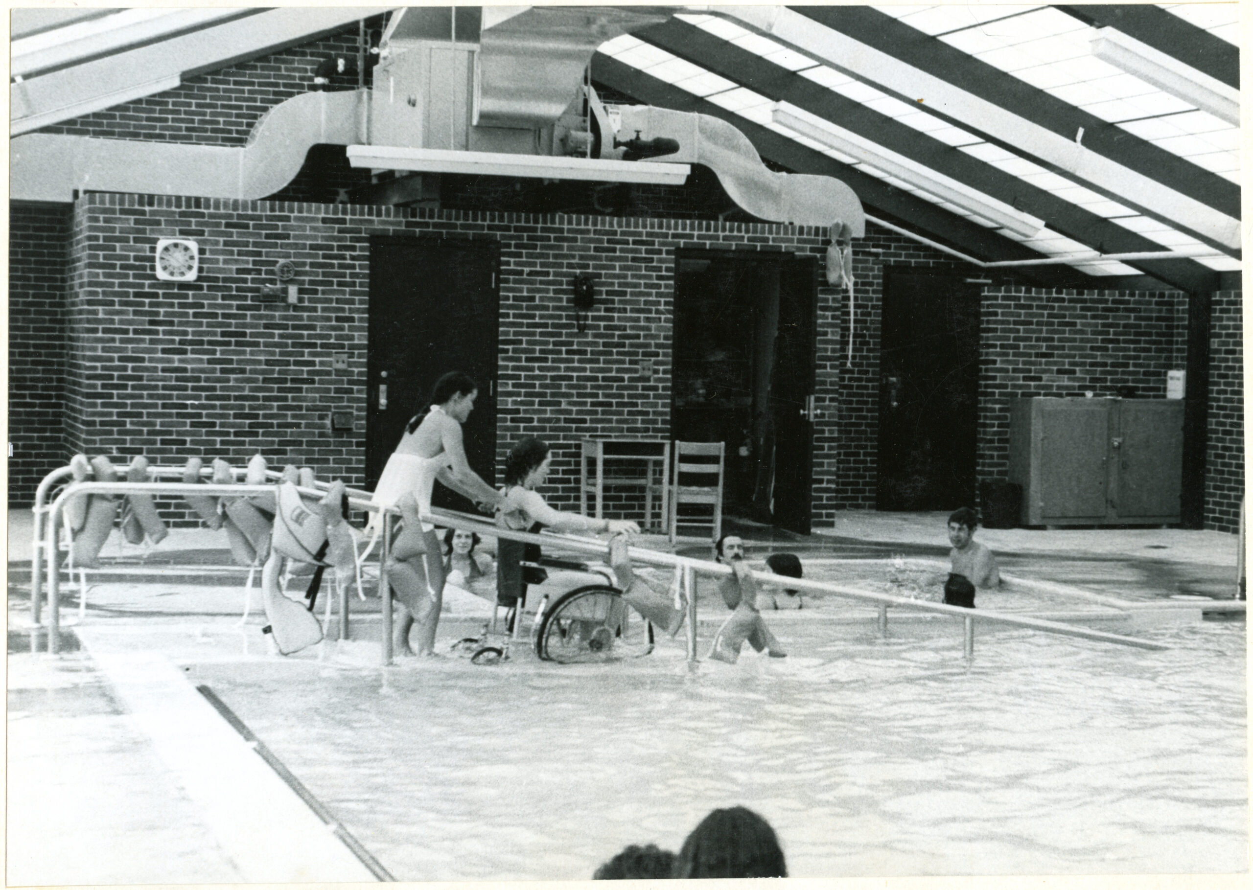 Black and white photo of an indoor swimming pool with a ramp that goes into it, and a woman pushing another woman into the pool in a wheelchair