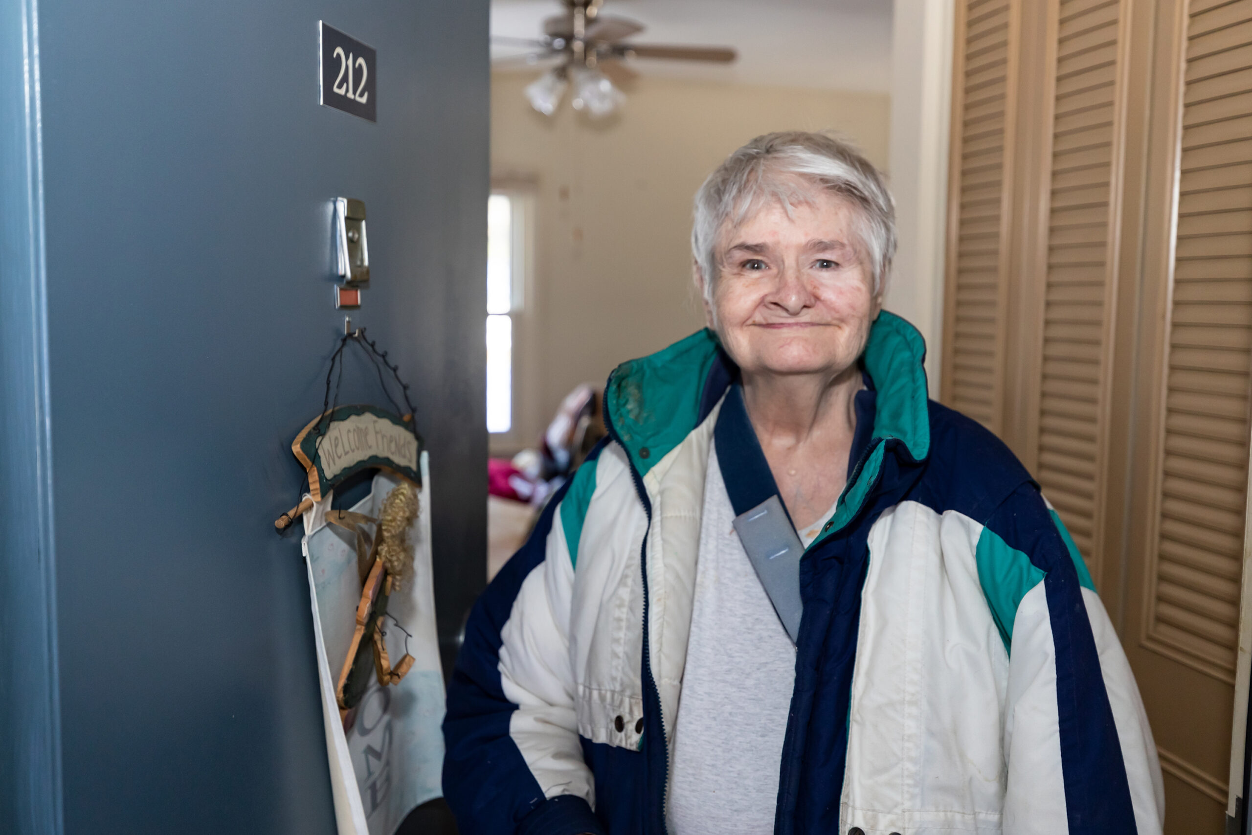 Maryann Preble stands just inside the open door of her apartment wearing a coat and smiling.