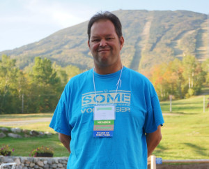 Eric stands wearing a bright blue shirt in front of a forested mountain