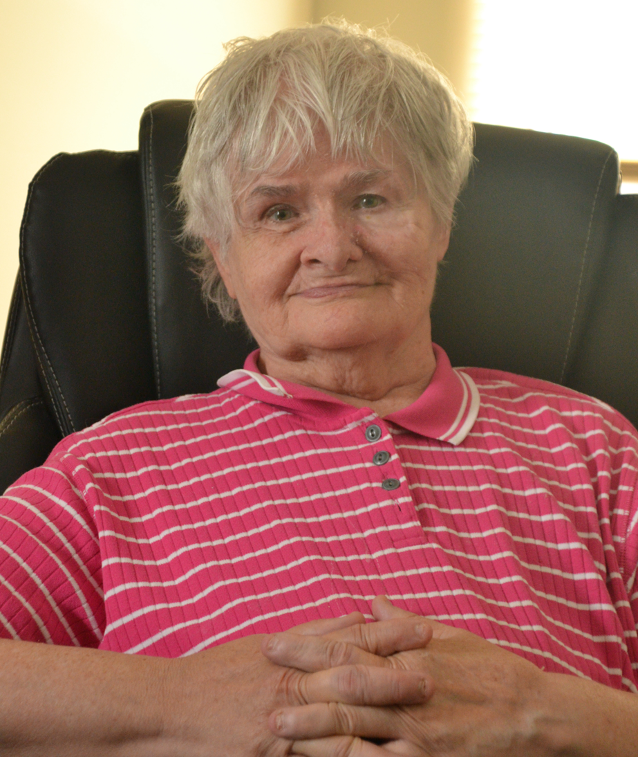 Maryann Preble sits in an easy chair smiling at the camera, arms folded over a pink and white striped shirt