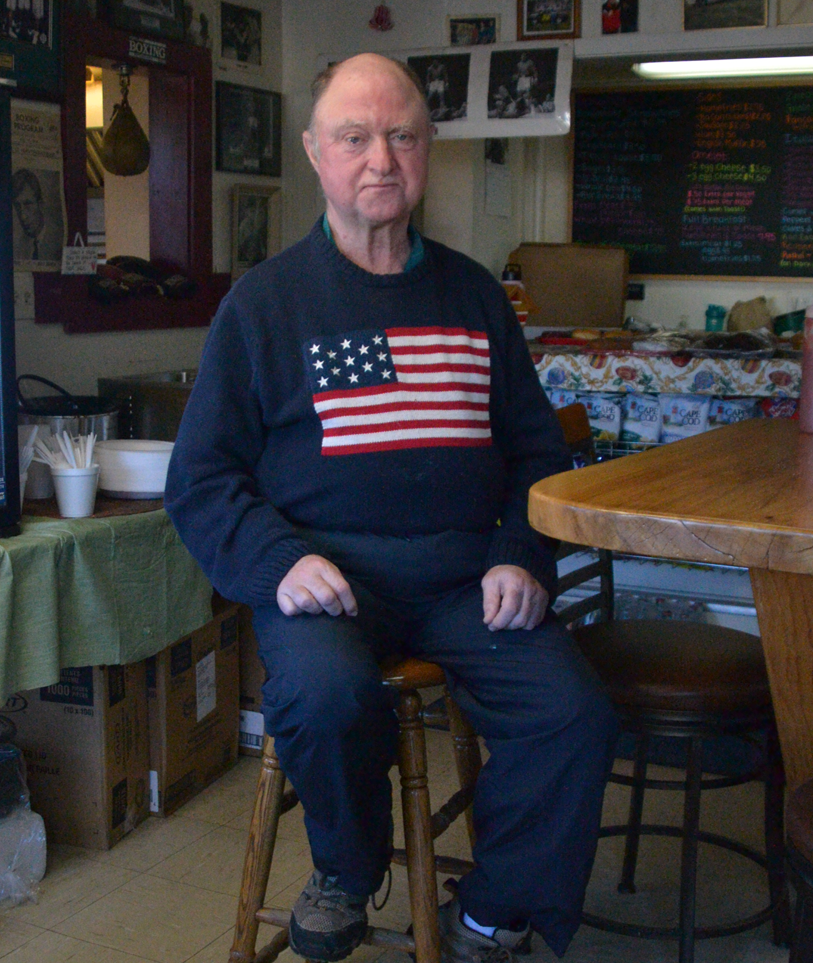 Paul Easton sits on a stool wearing a sweater with an American flag on it
