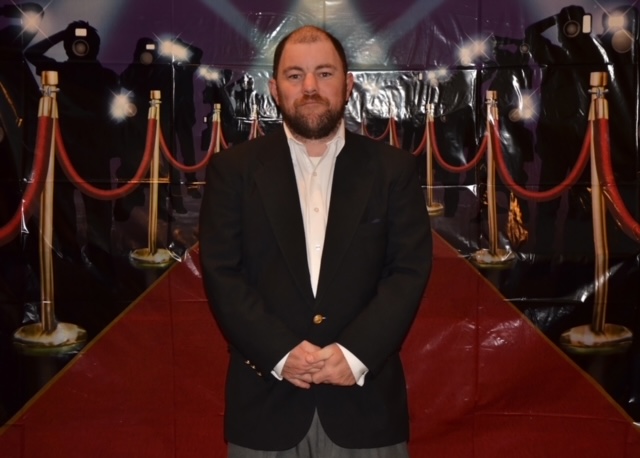 Jon McGovern, wearing a dark suit and white shirt, stands looking at the camera in front of a backdrop designed to look like a red carpet.